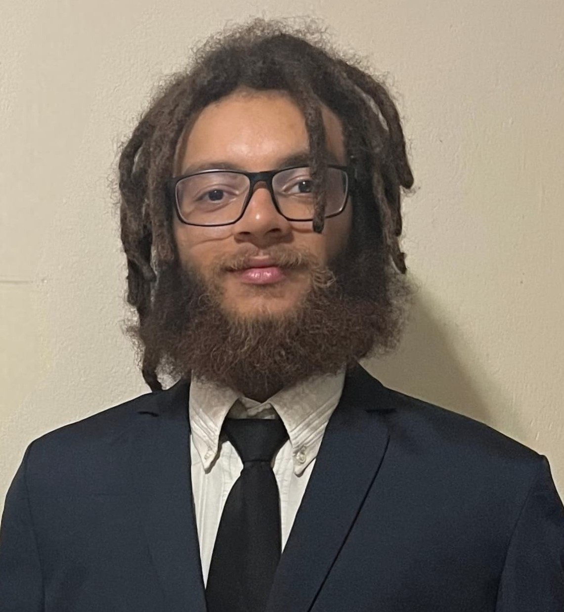 Rayan Trindade está com um leve sorriso, com cabelo castanho e barba. Ele está usando um terno preto.